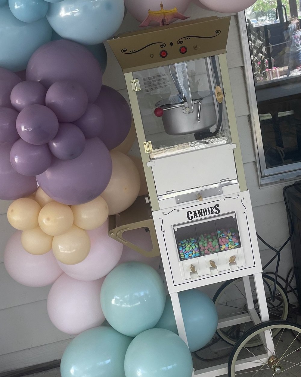 A popcorn machine on a white cart with a candy dispenser, surrounded by a pastel balloon garland in mint, pink and lilac