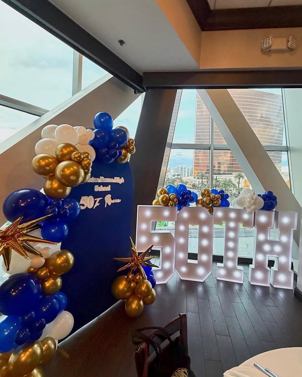 A high school 50th reunion backdrop in navy with gold and blue balloons and light-up letters, overlooking the Las Vegas Strip