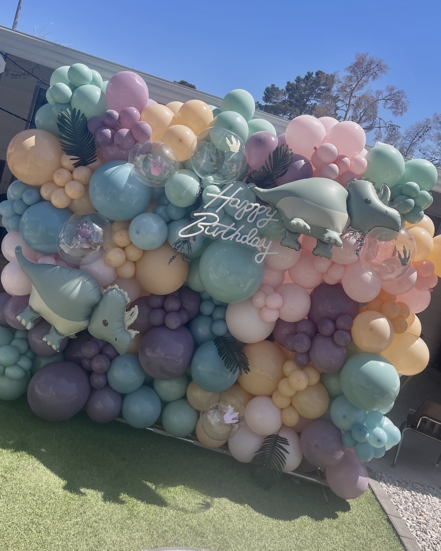 A full balloon wall in sage, dusty blue, cream and mauve with dinosaur balloons and a neon Happy Birthday sign