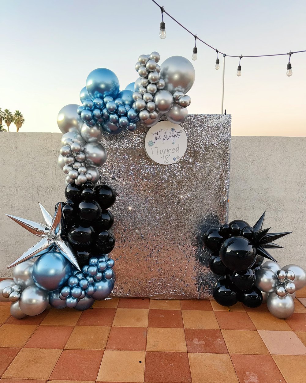 A square sequin backdrop with a silver, blue and black balloon garland, outdoors at sunset