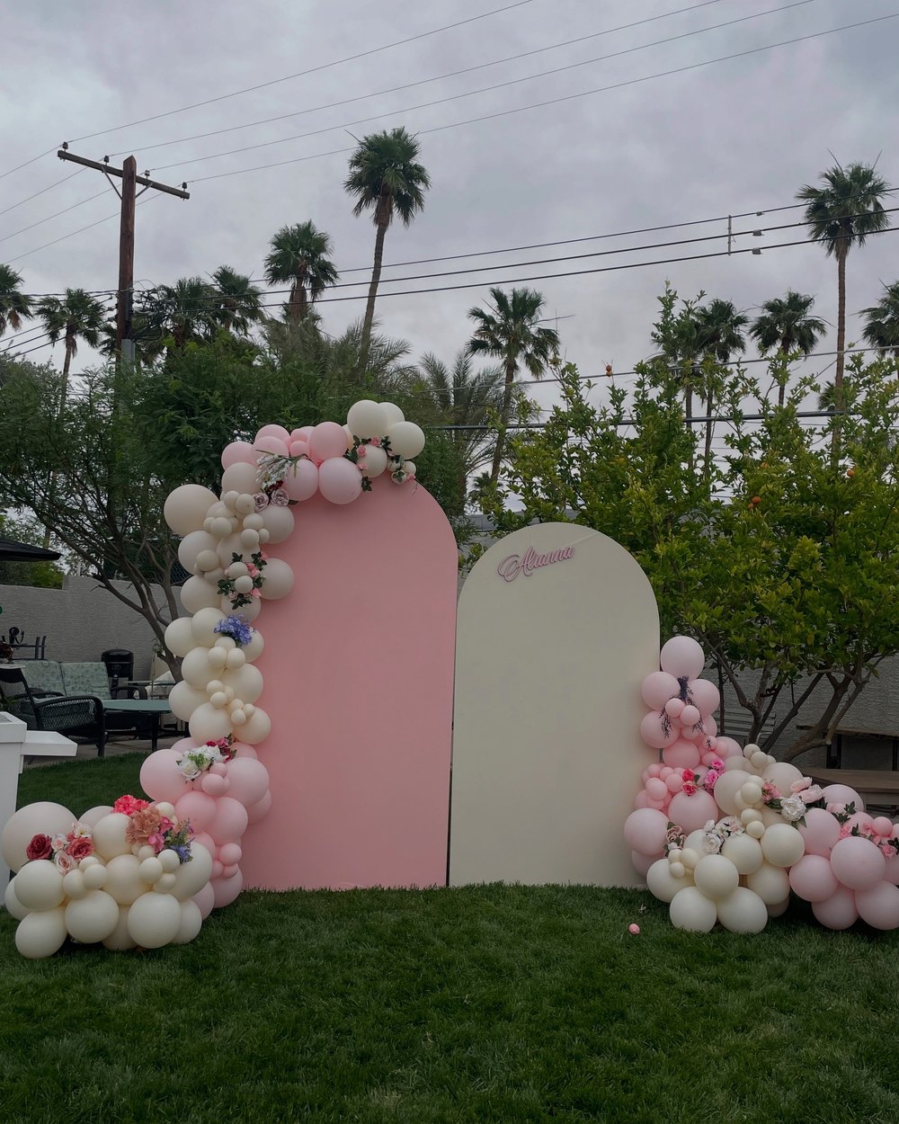 Two arched panels in pink and cream with cream and pink balloon garlands and flowers, on a lawn